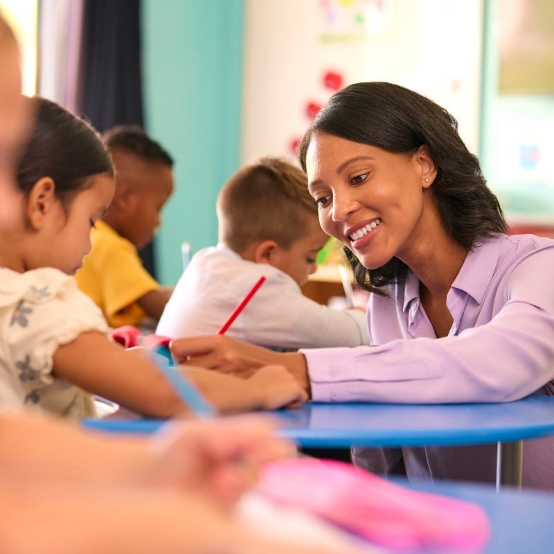 Female Primary Or Elementary School Teacher Helping Students At Desks In Multi-Cultural Class